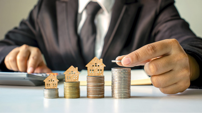 A person in a suit is stacking coins next to miniature house models, representing financial growth or real estate investment.