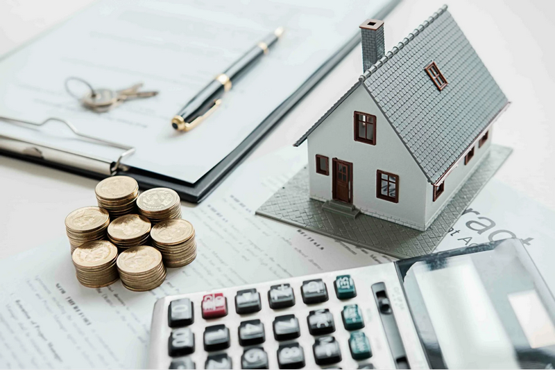 A small model house, stack of coins, calculator, pen, and keys on a document-filled desk.