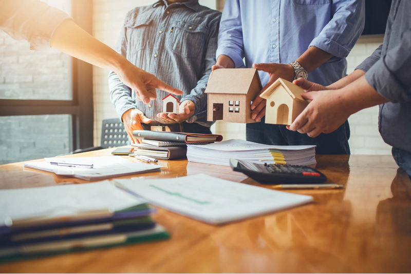 Three people holding model houses over a wooden table with documents and a calculator.