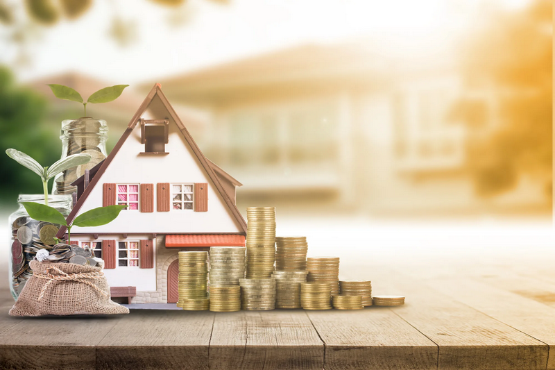 A model house is surrounded by stacks of coins, money bags, and jars with growing plants on a wooden surface.