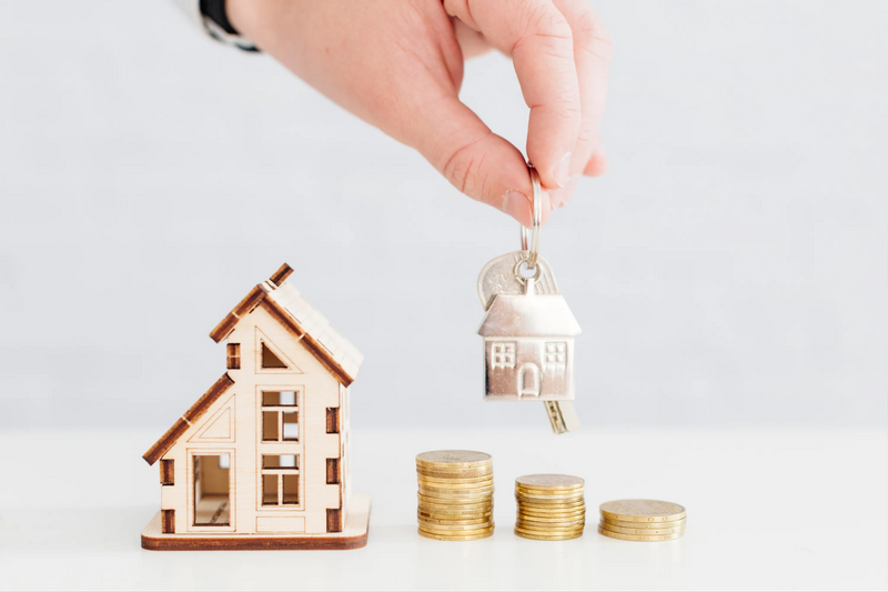 A hand holds a house key above a small wooden house model and stacked coins.