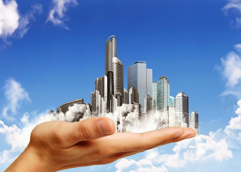 A hand holding a miniature city skyline surrounded by clouds against a blue sky.