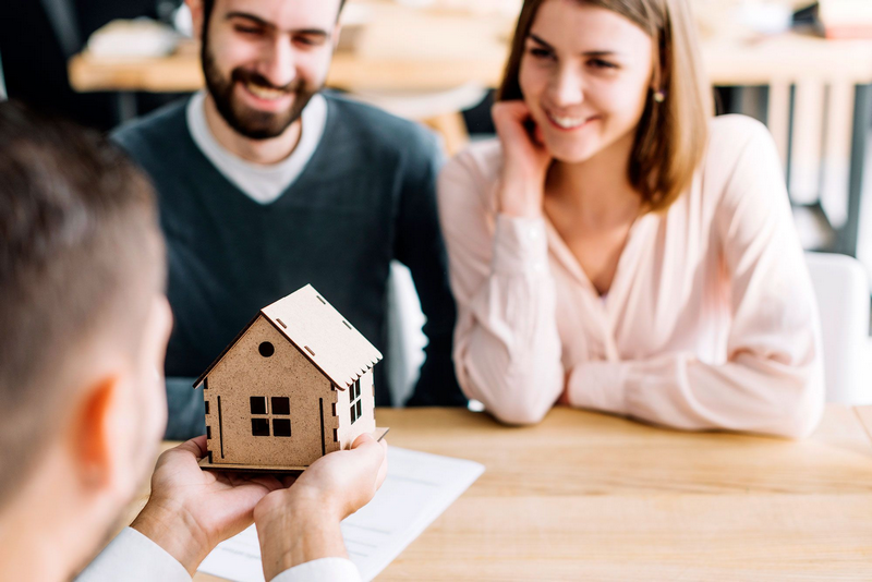 A couple sits across from a person holding a small wooden house model.